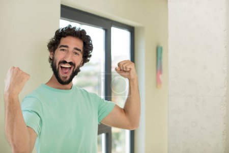 Téléchargez les photos : Jeune homme barbu fou se sentant heureux, satisfait et puissant, flexion ajustement et biceps musculaires, regardant fort après la salle de gym - en image libre de droit