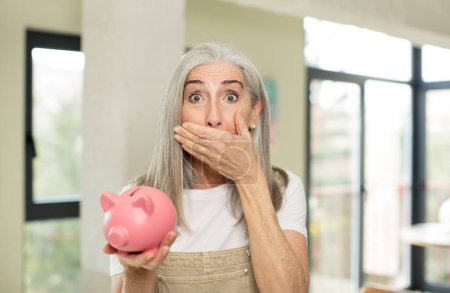 Photo for Pretty senior woman covering mouth with a hand and shocked or surprised expression. with a piggy bank - Royalty Free Image