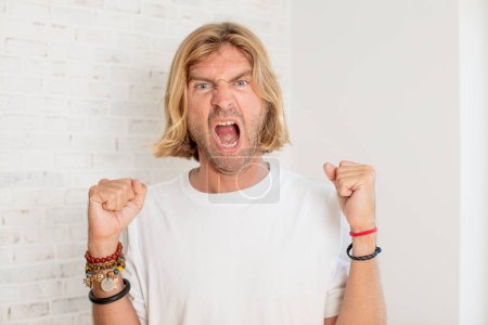 Photo for Young blond adult man shouting aggressively with an angry expression or with fists clenched celebrating success - Royalty Free Image