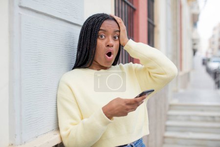 Photo for Black afro woman feeling extremely shocked and surprised. using a smartphone concept - Royalty Free Image