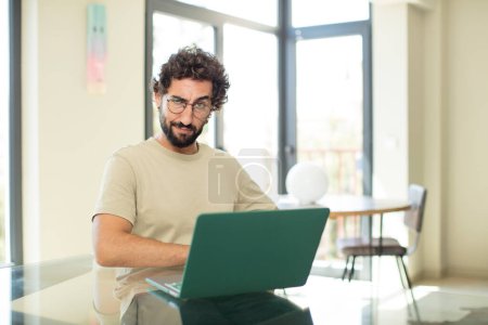 Foto de Joven hombre barbudo adulto con un ordenador portátil que mira orgulloso, confiado, fresco, descarado y arrogante, sintiéndose exitoso - Imagen libre de derechos