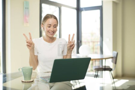 Téléchargez les photos : Caucasienne jolie femme avec un ordinateur portable souriant et regardant heureux, amical et satisfait, geste de victoire ou de paix avec les deux mains - en image libre de droit