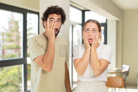 Téléchargez les photos : Jeune couple adulte se sentant choqué et effrayé, l'air ennuyé avec les mains sur les joues - en image libre de droit