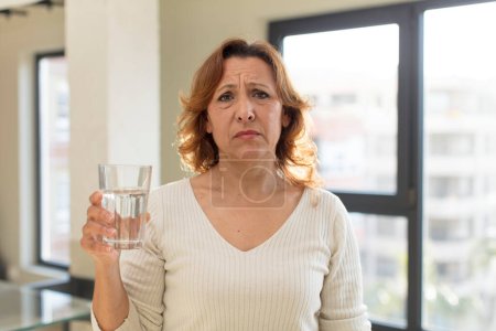 Téléchargez les photos : Jolie femme d'âge moyen se sentant triste et pleurnichard avec un regard malheureux et pleurer. verre d'eau - en image libre de droit