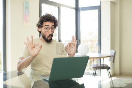 Photo for Young adult bearded man with a laptop feeling shocked, amazed and surprised, showing approval making okay sign with both hands - Royalty Free Image
