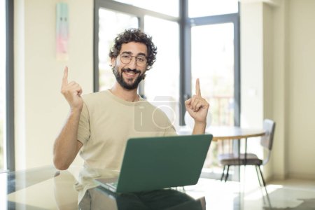 Photo for Young adult bearded man with a laptop feeling awed and open mouthed pointing upwards with a shocked and surprised look - Royalty Free Image