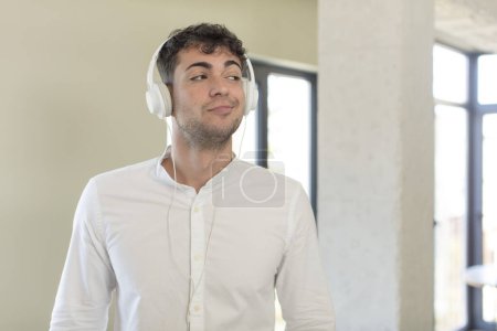 Téléchargez les photos : Jeune bel homme souriant et regardant avec une expression heureuse et confiante. écouter de la musique avec des écouteurs - en image libre de droit
