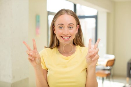 Photo for Pretty blond woman smiling and looking happy, friendly and satisfied, gesturing victory or peace with both hands - Royalty Free Image