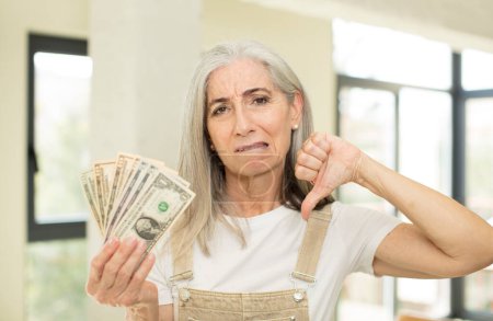 Foto de Mujer bastante mayor sintiéndose enfadada, mostrando los pulgares hacia abajo. con billetes en dólares - Imagen libre de derechos
