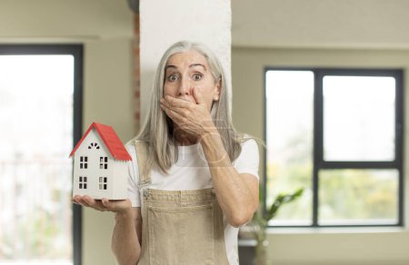 Photo for Pretty senior woman covering mouth with a hand and shocked or surprised expression. with a house model - Royalty Free Image
