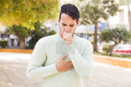 Téléchargez les photos : Jeune homme hispanique se sentant malade avec un mal de gorge et des symptômes de grippe, toux avec la bouche couverte - en image libre de droit