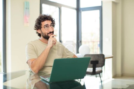 Téléchargez les photos : Jeune homme barbu adulte avec un ordinateur portable souriant joyeusement et rêvant ou doutant, regardant vers le côté - en image libre de droit