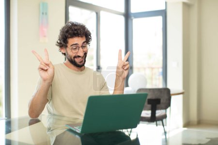 Foto de Joven hombre barbudo adulto con una computadora portátil sonriendo y mirando feliz, amistoso y satisfecho, haciendo un gesto de victoria o paz con ambas manos - Imagen libre de derechos