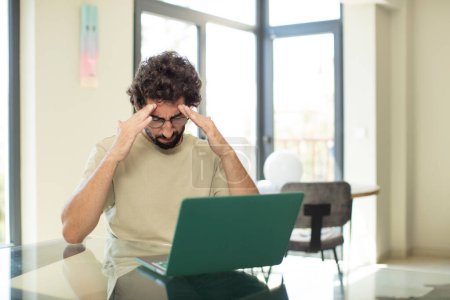 Photo for Young adult bearded man with a laptop looking concentrated, thoughtful and inspired, brainstorming and imagining with hands on forehead - Royalty Free Image