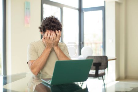 Photo for Young adult bearded man with a laptop feeling sad, frustrated, nervous and depressed, covering face with both hands, crying - Royalty Free Image