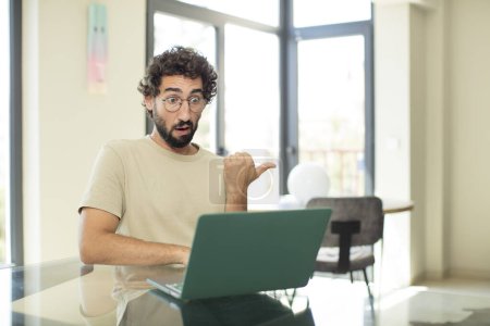 Photo for Young adult bearded man with a laptop looking astonished in disbelief, pointing at object on the side and saying wow, unbelievable - Royalty Free Image