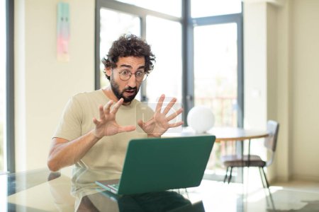 Foto de Joven hombre barbudo adulto con un ordenador portátil sentirse aterrorizado, retroceder y gritar de horror y pánico, reaccionando a una pesadilla - Imagen libre de derechos