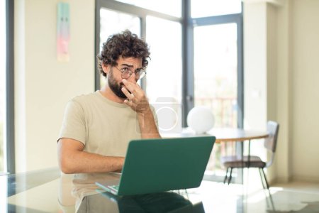 Téléchargez les photos : Jeune homme barbu adulte avec un ordinateur portable se sentant dégoûté, tenant le nez pour éviter de sentir une puanteur sale et désagréable - en image libre de droit