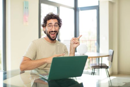 Foto de Joven hombre barbudo adulto con una computadora portátil sonriendo alegremente y apuntando a copiar espacio en la palma de la mano, mostrando o publicitando un objeto - Imagen libre de derechos