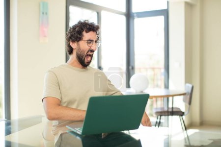 Photo for Young adult bearded man with a laptop shouting aggressively, looking very angry, frustrated, outraged or annoyed, screaming no - Royalty Free Image