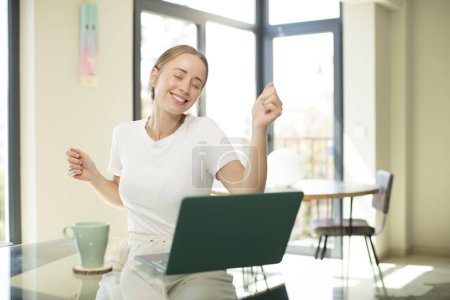 Foto de Mujer guapa caucásica con una laptop sonriendo, sintiéndose despreocupada, relajada y feliz, bailando y escuchando música, divirtiéndose en una fiesta - Imagen libre de derechos