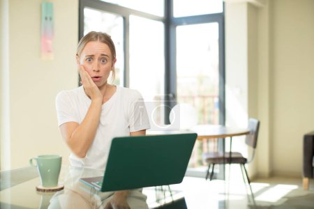 Téléchargez les photos : Caucasienne jolie femme avec un ordinateur portable se sentant choqué et étonné tenant face à la main dans l'incrédulité avec la bouche grande ouverte - en image libre de droit