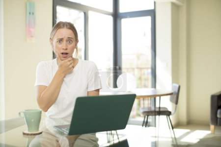 Photo for Caucasian pretty woman with a laptop with mouth and eyes wide open and hand on chin, feeling unpleasantly shocked, saying what or wow - Royalty Free Image