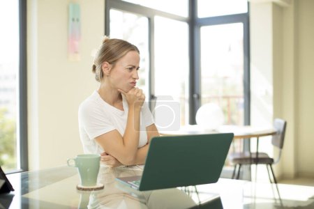 Téléchargez les photos : Caucasienne jolie femme avec un ordinateur portable se sentant confus ou douteux, se concentrant sur une idée, réfléchissant dur, cherchant à copier l'espace sur le côté - en image libre de droit