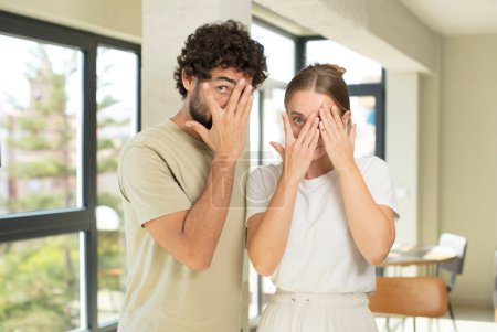 Photo for Young adult couple covering face with hands, peeking between fingers with surprised expression and looking to the side - Royalty Free Image