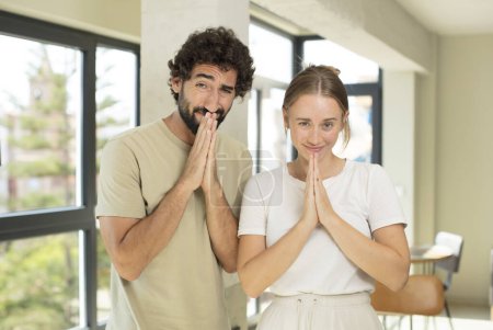 Photo for Young adult couple feeling worried, hopeful and religious, praying faithfully with palms pressed, begging forgiveness - Royalty Free Image