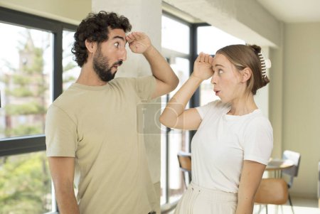 Photo for Young adult couple looking bewildered and astonished, with hand over forehead looking far away, watching or searching - Royalty Free Image
