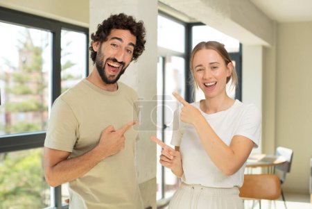 Photo for Young adult couple smiling cheerfully and pointing to copy space on palm on the side, showing or advertising an object - Royalty Free Image