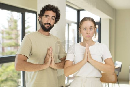 Photo for Young adult couple feeling worried, hopeful and religious, praying faithfully with palms pressed, begging forgiveness - Royalty Free Image