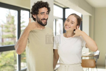 Foto de Joven pareja de adultos sonriendo alegremente y señalando a la cámara mientras que hacer una llamada más tarde gesto, hablando por teléfono - Imagen libre de derechos