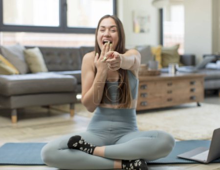Photo for Young adult woman practicing yoga laughing at you, pointing to camera and making fun of or mocking you - Royalty Free Image