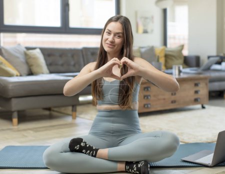 Téléchargez les photos : Jeune femme adulte pratiquant le yoga souriant et se sentant heureux, mignon, romantique et amoureux, ce qui rend la forme du cœur avec les deux mains - en image libre de droit