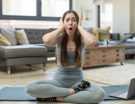 Téléchargez les photos : Jeune femme adulte pratiquant le yoga à l'air excité et surpris, bouche ouverte avec les deux mains sur la tête, se sentant comme un gagnant chanceux - en image libre de droit