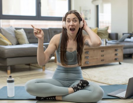 Foto de Joven mujer adulta practicando yoga riéndose, luciendo feliz, positiva y sorprendida, realizando una gran idea apuntando al espacio lateral de copia - Imagen libre de derechos