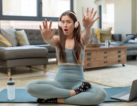 Foto de Mujer adulta joven practicando yoga sintiéndose estupefacta y asustada, temiendo algo aterrador, con las manos abiertas delante diciendo aléjate - Imagen libre de derechos