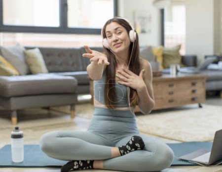 Téléchargez les photos : Jeune femme adulte pratiquant le yoga se sentant heureuse et amoureuse, souriant d'une main à côté du cœur et l'autre étirée à l'avant - en image libre de droit
