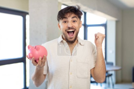 Photo for Young handsome man feeling shocked,laughing and celebrating success. piggy bank concept - Royalty Free Image