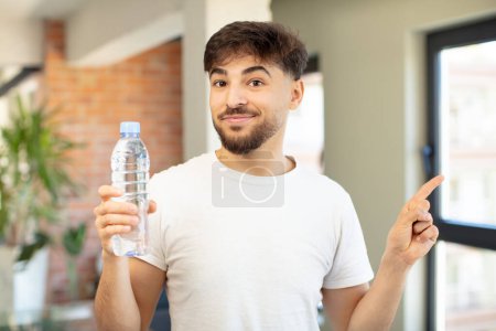 Téléchargez les photos : Jeune bel homme souriant joyeusement, se sentant heureux et pointant vers le côté. concept de bouteille d'eau - en image libre de droit