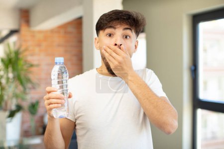 Téléchargez les photos : Jeune homme beau couvrant la bouche avec une main et une expression choquée ou surprise. concept de bouteille d'eau - en image libre de droit