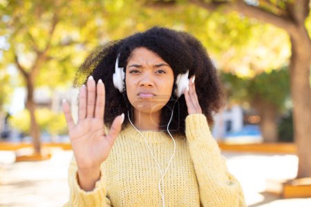 Foto de Bastante afro negro mujer mirando serio mostrando abierta palma haciendo gesto de parada. escuchar música con auriculares - Imagen libre de derechos