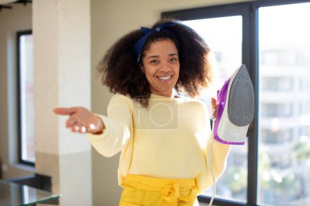 Foto de Mujer negra bastante afro sonriendo felizmente y ofreciendo o mostrando un concepto. ropa de hierro concepto de lavandería - Imagen libre de derechos