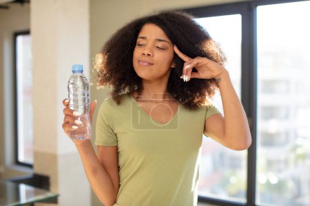 Foto de Mujer negra bastante afro mirando sorprendida, realizando un nuevo pensamiento, idea o concepto. concepto de botella de agua - Imagen libre de derechos