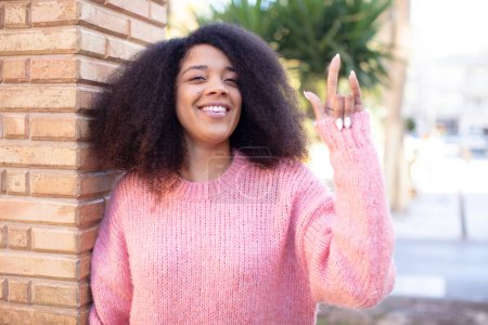 Foto de Mujer bonita afroamericana sintiéndose feliz, divertida, confiada, positiva y rebelde, haciendo el cartel del rock o del heavy metal con la mano - Imagen libre de derechos
