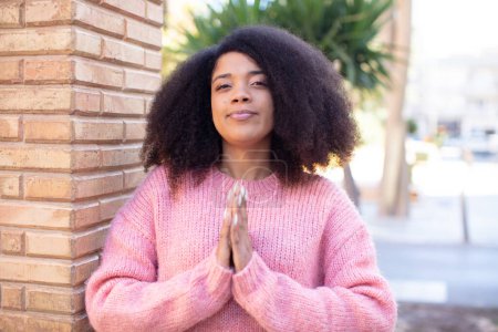 Téléchargez les photos : Afro-américaine jolie femme se sentant inquiète, pleine d'espoir et religieuse, priant fidèlement avec les paumes pressées, implorant pardon - en image libre de droit