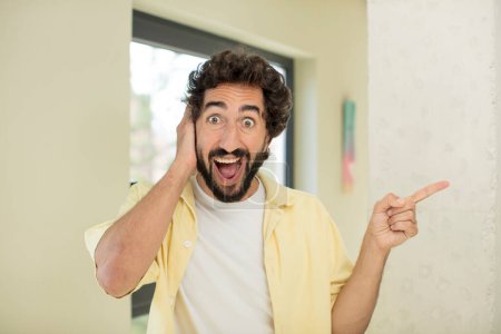 Téléchargez les photos : Jeune homme barbu fou riant, regardant heureux, positif et surpris, réalisant une grande idée pointant vers l'espace de copie latérale - en image libre de droit