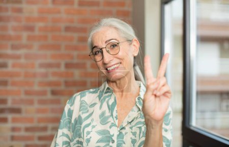 Foto de Anciana bonita mujer sonriendo y luciendo feliz, despreocupada y positiva, haciendo gestos de victoria o paz con una mano - Imagen libre de derechos